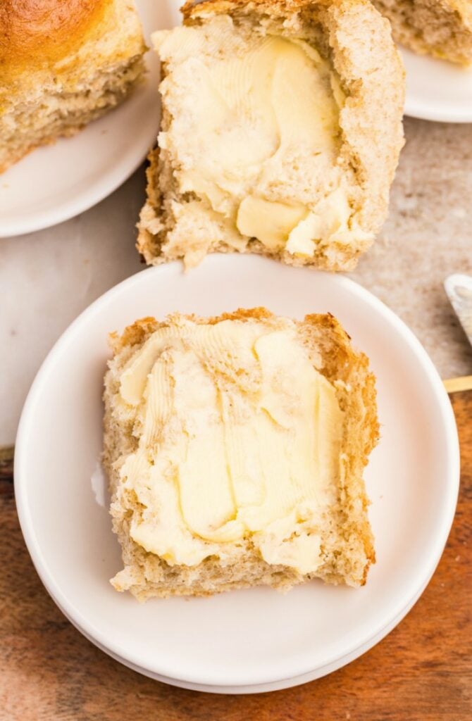 A piece of bread roll topped with butter on a white plate, with another buttered roll partially visible above.