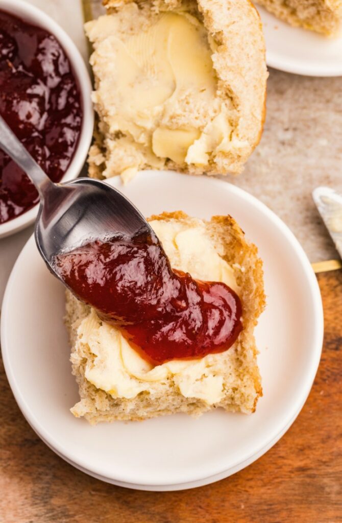 A spoon spreads red jam onto a buttered biscuit half on a white plate, with a bowl of jam and another biscuit in the background.