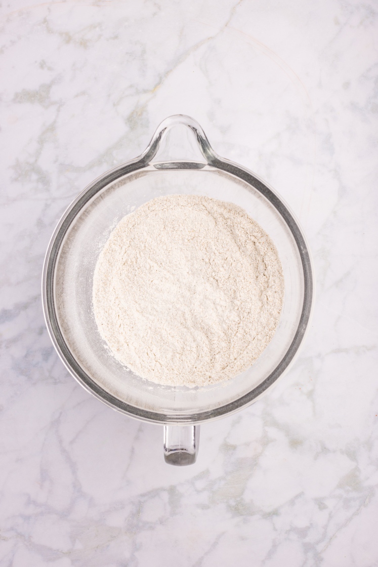 Glass mixing bowl filled with combined dry ingredients for rye dinner rolls.