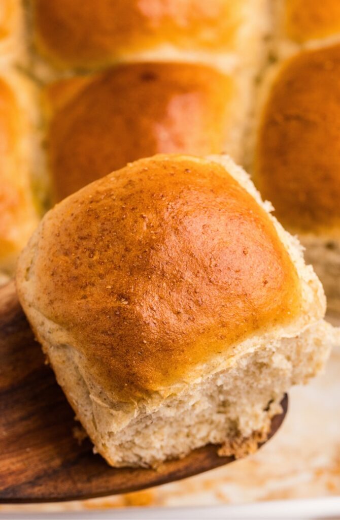 A golden-brown dinner roll being lifted from a tray of freshly baked rolls with a wooden spatula.