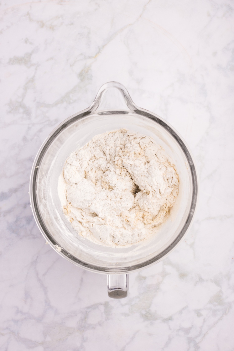 Glass mixing bowl with dough that has just started to come together, showing a rough and floury texture.