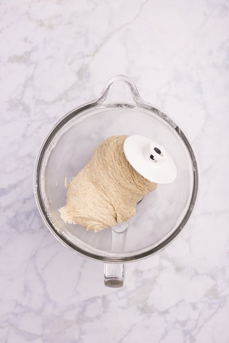 Stand mixer bowl with kneaded rye dinner roll dough wrapped around the dough hook attachment.