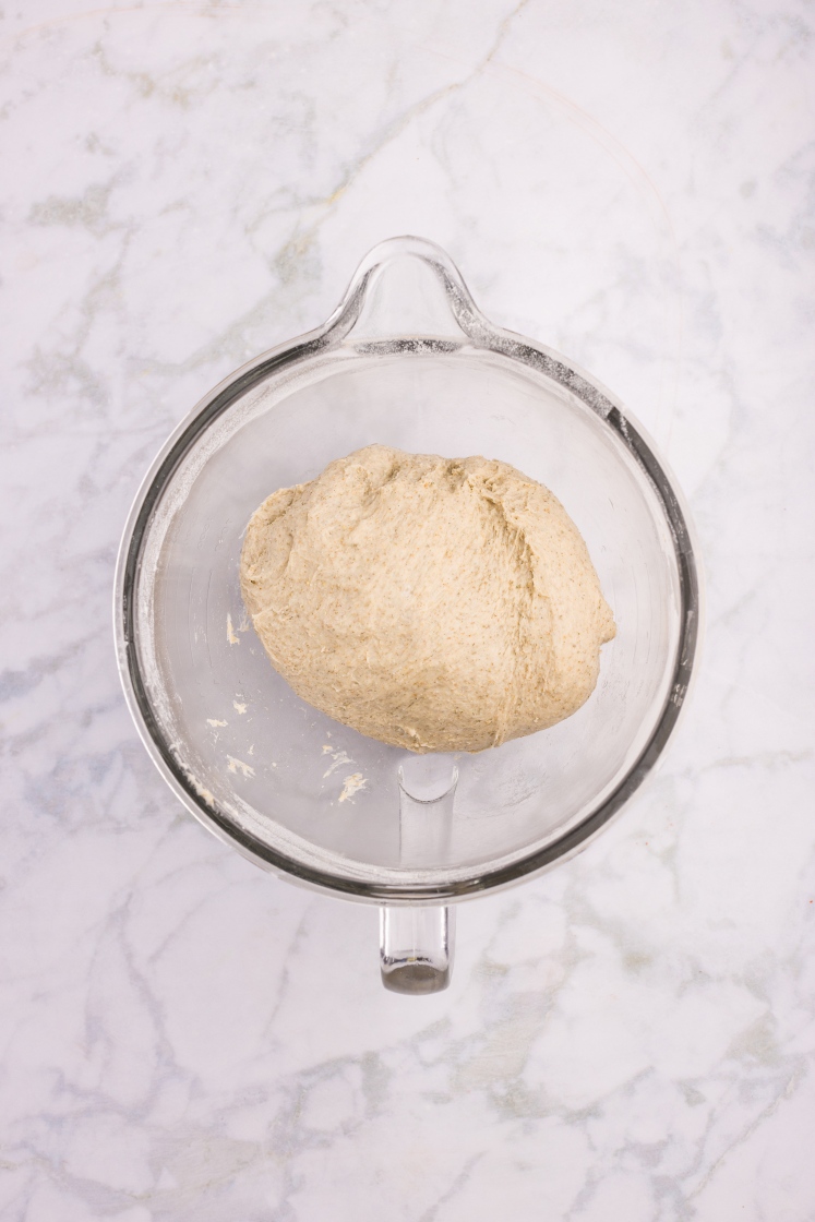 Ball of rye dinner roll dough resting in a clear mixing bowl on a marble surface.