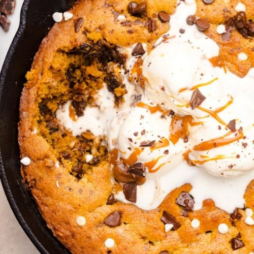 A skillet cookie topped with three scoops of vanilla ice cream, chocolate chips, caramel sauce, and chocolate chunks, with a portion missing.