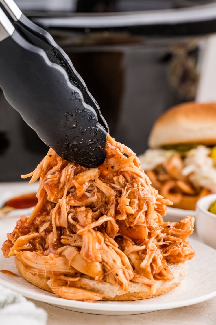 A pair of tongs places shredded barbecue chicken onto a sandwich bun, with a slow cooker and another sandwich visible in the background.