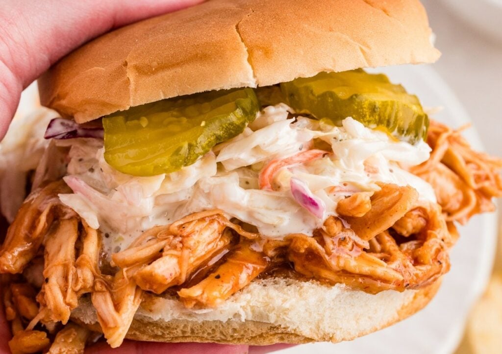 A hand holds a sandwich with shredded chicken, coleslaw, and pickle slices in a hamburger bun.