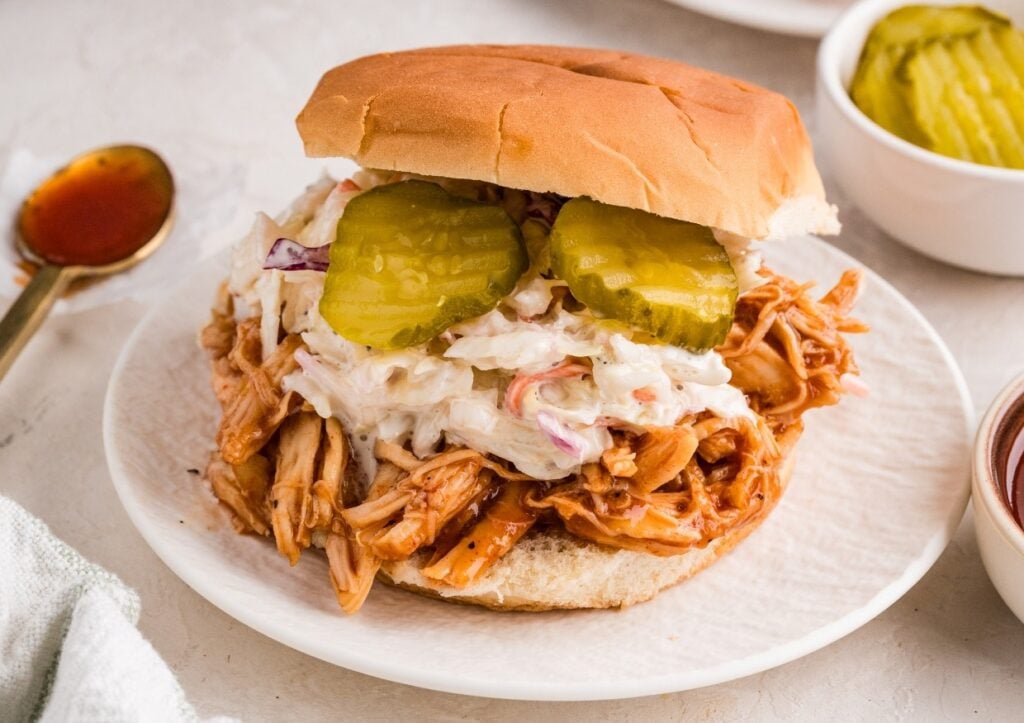 Shredded barbecue chicken sandwich topped with coleslaw and pickle slices on a bun, served on a white plate with bowls of pickles and sauce nearby.