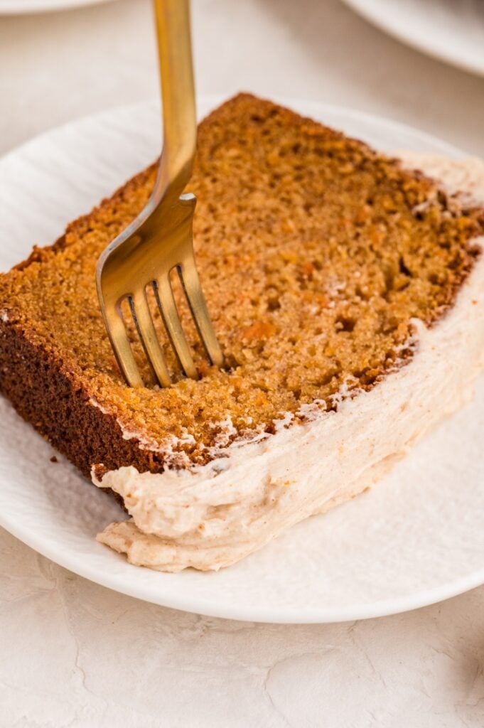 A gold fork slices into a piece of frosted carrot cake on a white plate.
