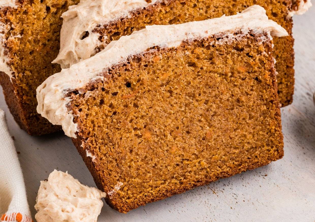 A close-up of a slice of pumpkin bread topped with a layer of creamy frosting, resting on a light-colored surface.
