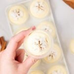 A hand holds a small cup of vanilla RumChata pudding shot above a tray of similar pudding shots, each topped with whipped cream and a sprinkle of spice.