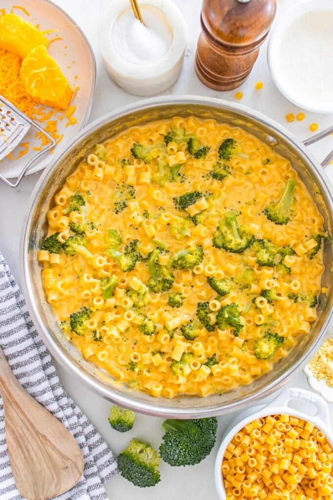 Creamy broccoli cheddar ditalini pasta in a skillet, surrounded by shredded cheese, pasta, broccoli florets, and seasonings on the counter.