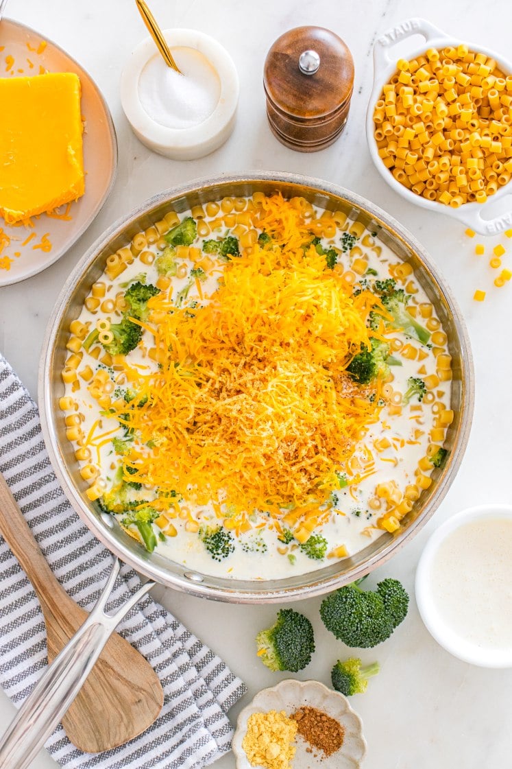 Skillet filled with ditalini pasta, broccoli, cream, and a large pile of shredded cheddar cheese before mixing to make broccoli cheddar pasta.