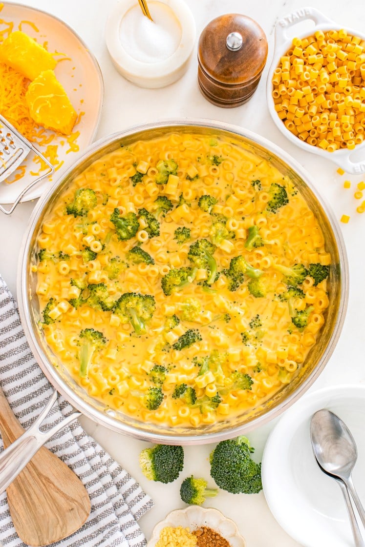 Creamy broccoli cheddar ditalini pasta simmering in a skillet, with shredded cheese, pasta, spices, and serving dishes arranged around it.