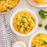 Bowls of creamy broccoli cheddar ditalini pasta on a countertop, with extra pasta, broccoli, shredded cheese, and seasonings nearby.