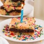 A square piece of sprinkled birthday cake with a lit blue candle in the center, served on a white plate with colorful sprinkles.