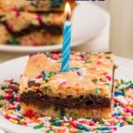 A brownie topped with colorful sprinkles and a lit blue birthday candle sits on a plate, with more brownies stacked in the background.