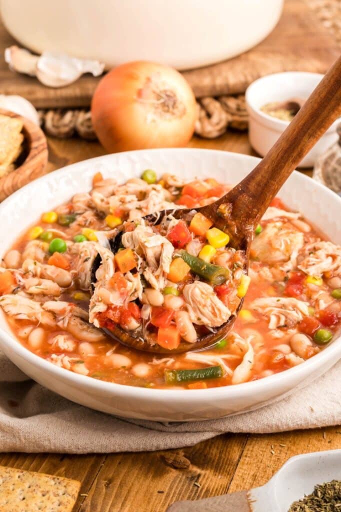 A wooden ladle scoops chicken and vegetable soup with beans, corn, carrots, peas, and tomatoes from a white bowl on a rustic table.