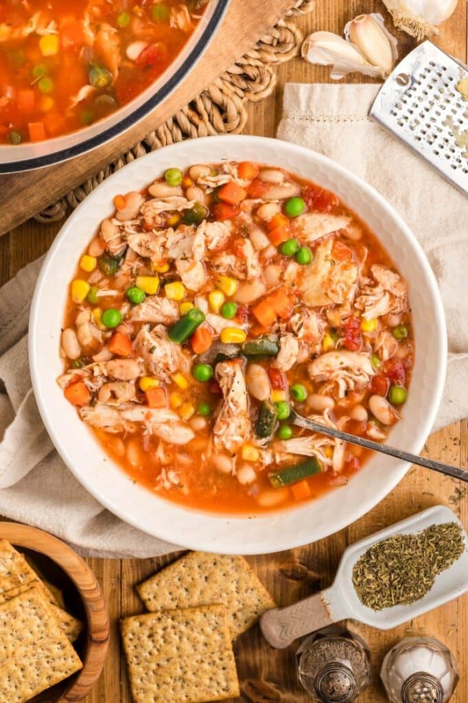 A bowl of chicken and vegetable soup with beans, corn, peas, carrots, and tomatoes, served with a spoon and crackers on the side.