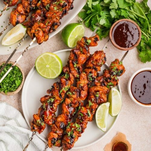 Chipotle chicken skewers on plates with lime wedges, cilantro, and dipping sauce.