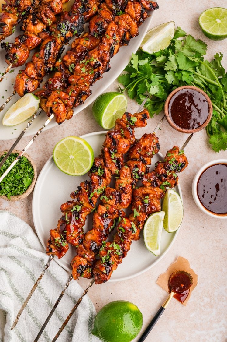 Chipotle chicken skewers on plates with lime wedges, cilantro, and dipping sauce.