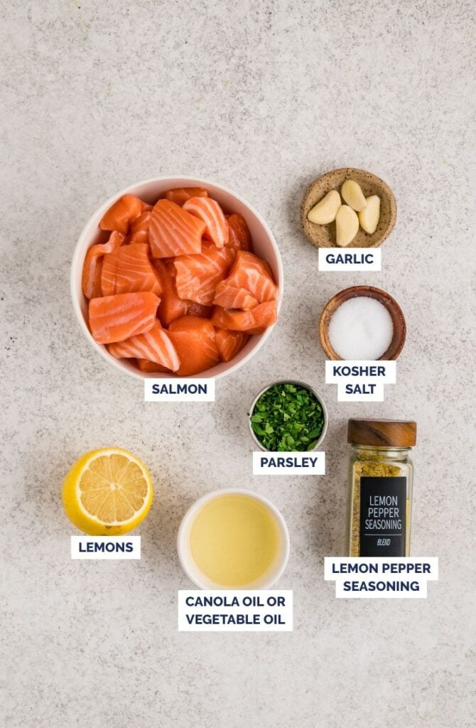 A simple ingredient lineup for lemon garlic salmon skewers: cubed salmon, fresh garlic, lemon, olive oil, chopped herbs, salt, and lemon pepper seasoning laid out and ready to prep.