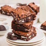 A stack of fudgy peppermint patty brownies on a white plate, showing layers of rich chocolate brownie and creamy mint filling, with chocolate patties on the side.