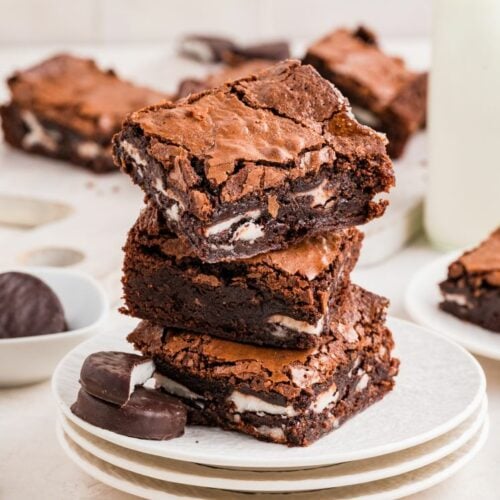 A stack of fudgy peppermint patty brownies on a white plate, showing layers of rich chocolate brownie and creamy mint filling, with chocolate patties on the side.