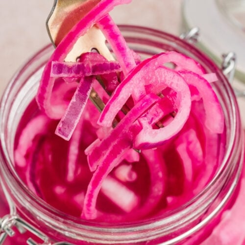 Close-up of a gold fork lifting a tangle of dripping, bright pink pickled red onion strands out of a clear glass jar.