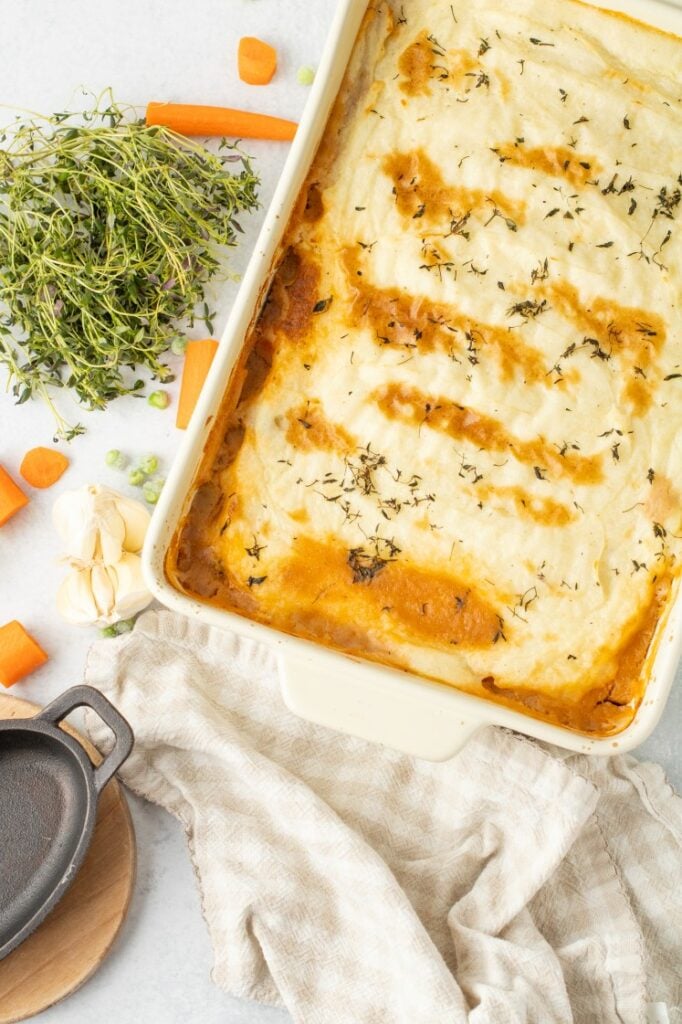 A golden-topped shepherd’s pie fresh from the oven, with creamy mashed potatoes lightly browned and sprinkled with herbs, surrounded by carrots, garlic, and fresh thyme for a cozy, classic feel.