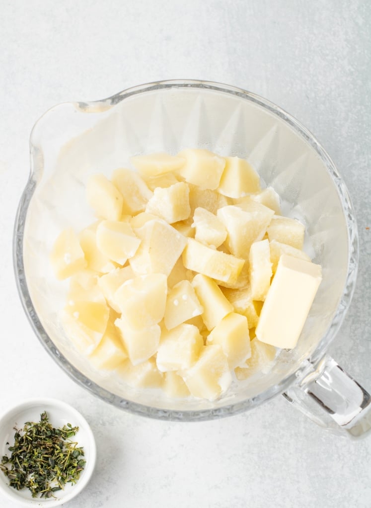 The cooked potatoes are transferred to a bowl while still hot, with butter added on top so it can melt easily before mashing. This sets them up to become smooth and creamy for the shepherd’s pie topping.