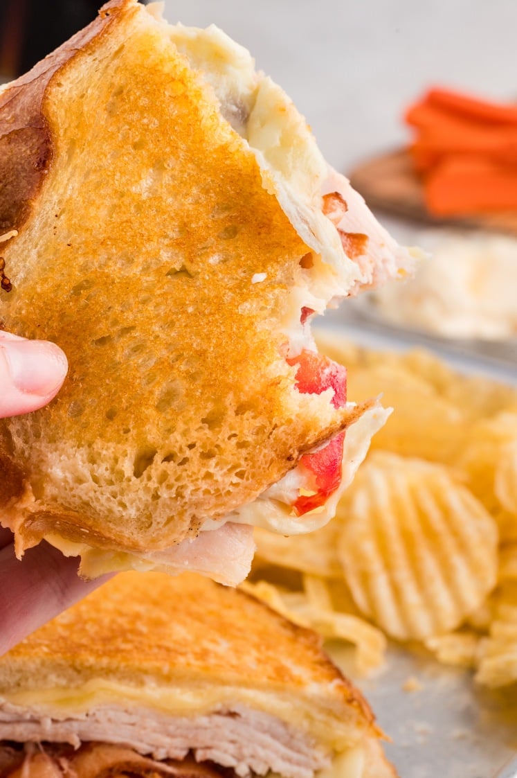 A hand holds a grilled sandwich with a bite taken out, revealing turkey, cheese, and tomato; potato chips are visible in the background.