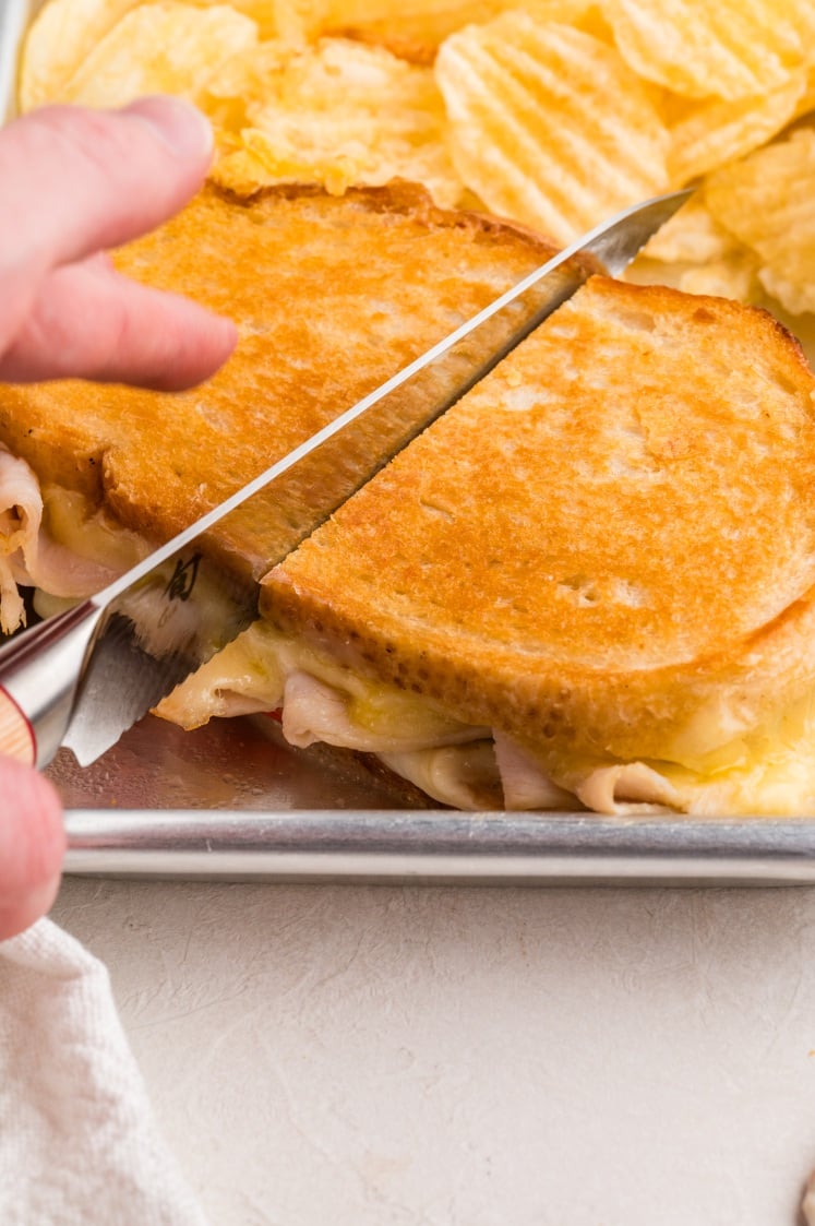 A hand slices a grilled cheese and turkey sandwich on a metal tray with a side of potato chips.