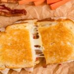 A grilled cheese sandwich cut in half with melted cheese stretching, served with bacon, carrot sticks, and potato chips on brown parchment paper.