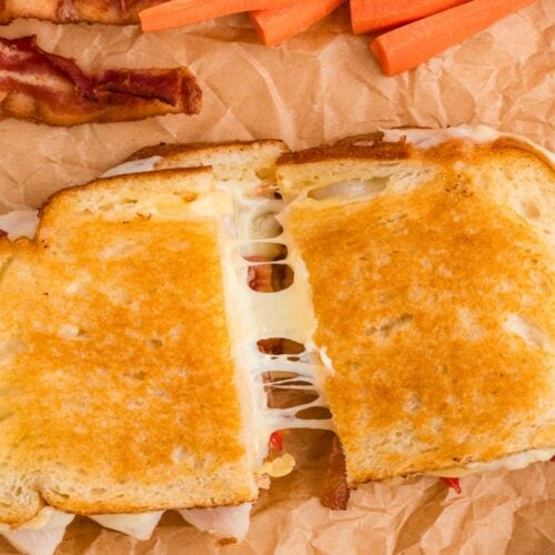 A grilled cheese sandwich cut in half with melted cheese stretching, served with bacon, carrot sticks, and potato chips on brown parchment paper.