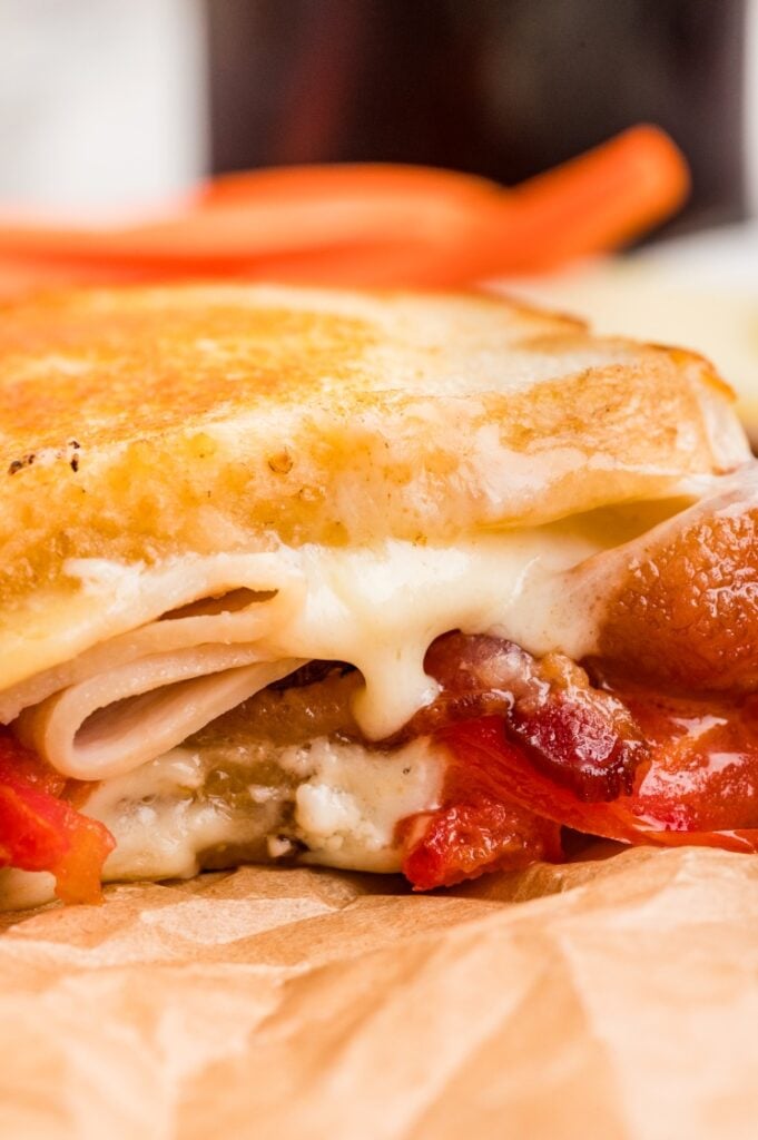 Close-up of a grilled cheese sandwich with melted cheese, turkey, bacon, and tomato on toasted bread, resting on brown parchment paper.
