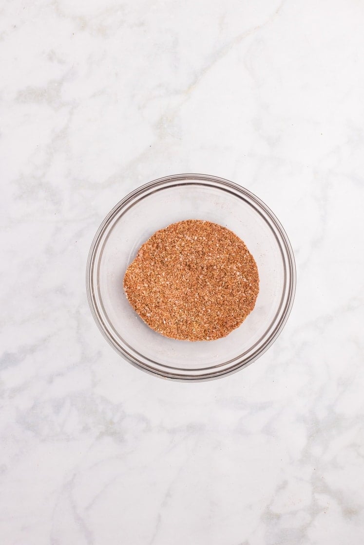 Small glass bowl filled with brown spice rub seasoning on a marble surface.