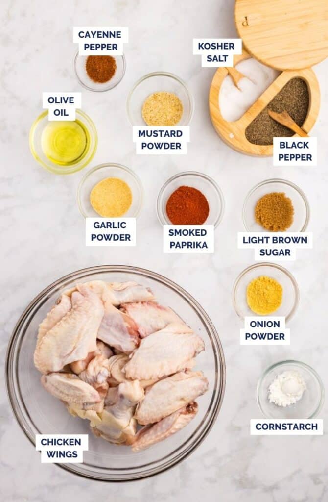 Raw chicken wings with small bowls of oil, spices, seasonings, and cornstarch laid out on a marble surface.