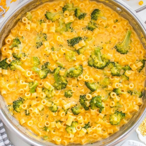 Overhead view of a large skillet of broccoli cheddar ditalini surrounded by ingredients including shredded cheddar, dry pasta, and broccoli florets.