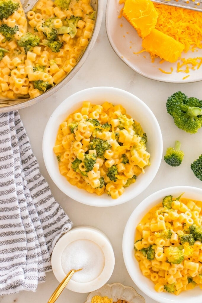 Overhead view of two white bowls of broccoli cheddar ditalini next to a skillet, a block of cheddar, and fresh broccoli florets.