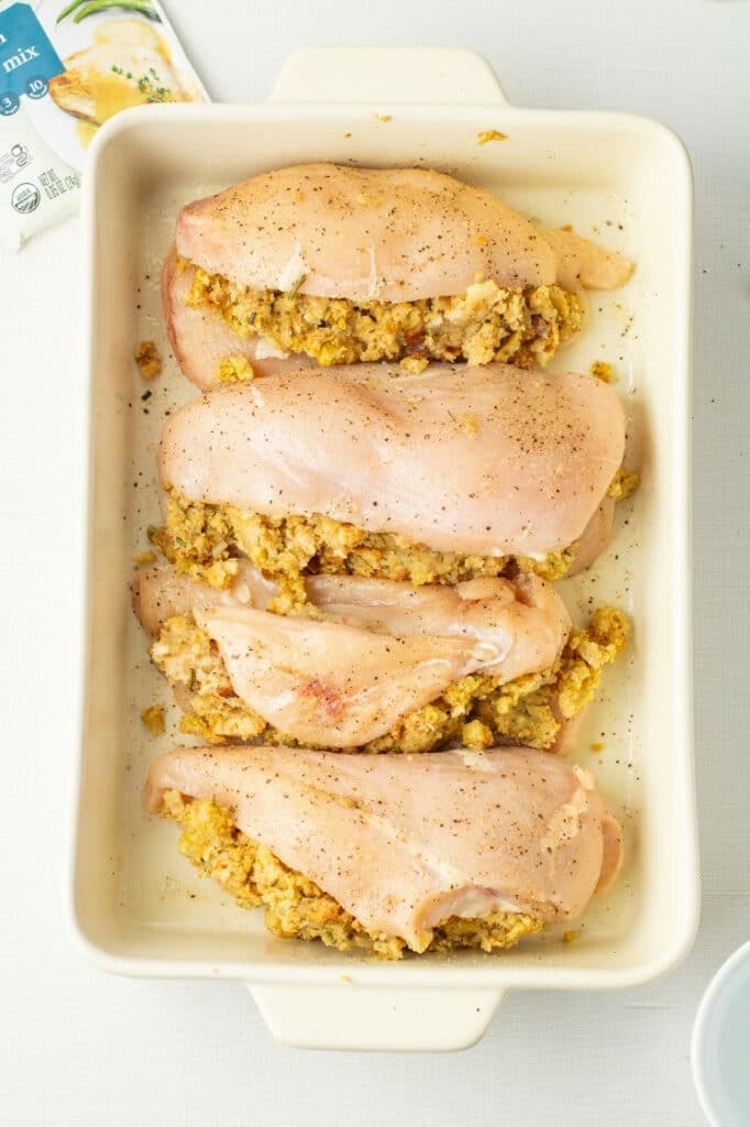 Four raw seasoned chicken breasts stuffed with cornbread stuffing in a white baking dish, uncooked.