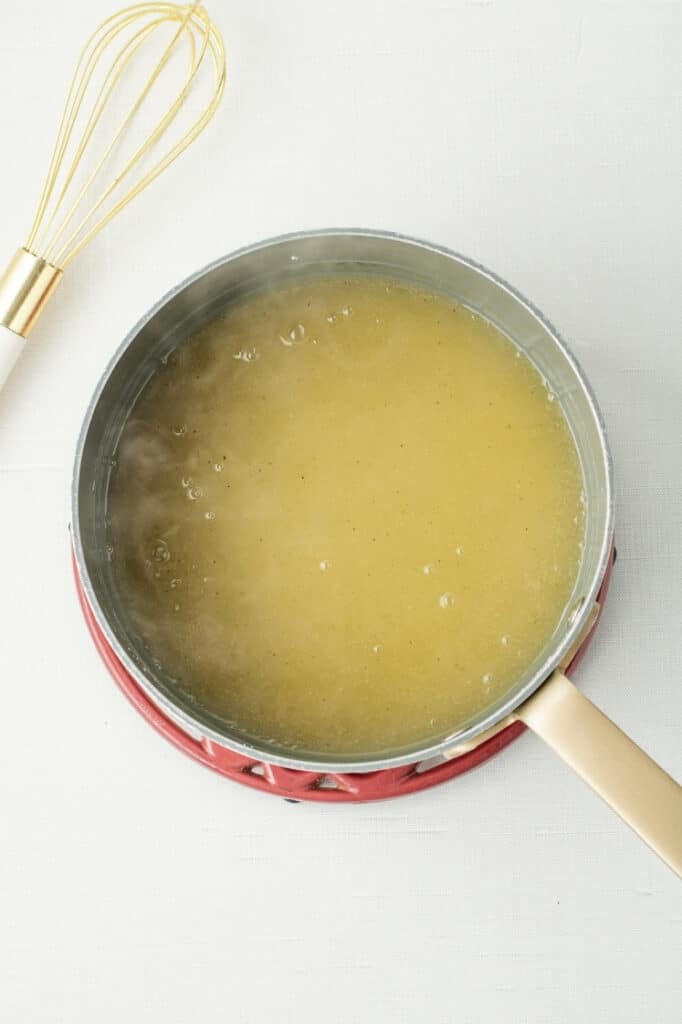 Overhead view of golden chicken broth in a saucepan on a trivet, with a whisk nearby.
