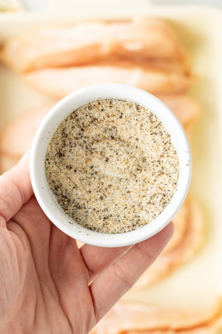 A hand holding a small white ramekin of salt and pepper seasoning, with raw chicken in the background.
