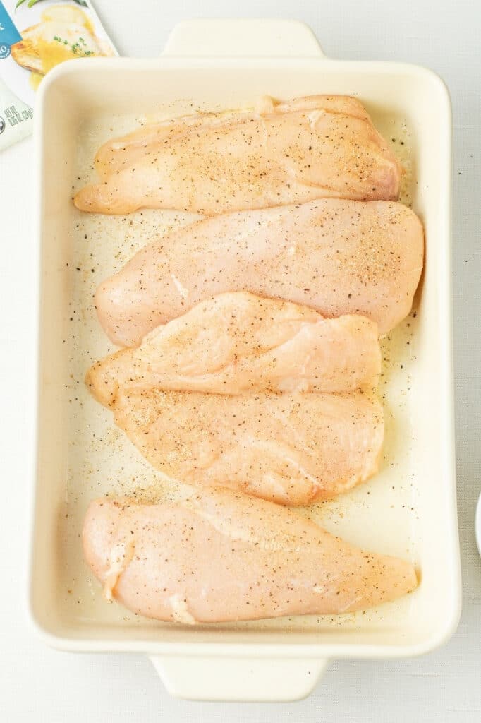 Four seasoned raw chicken breasts laid flat in a white rectangular baking dish.