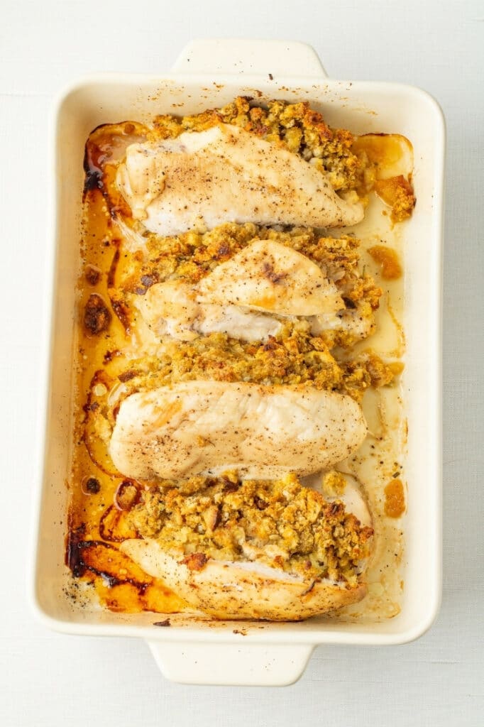 Four baked stuffed chicken breasts in a white baking dish, golden and juicy with herb stuffing visible.