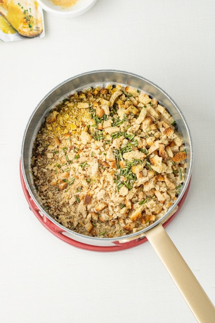 A saucepan filled with dry bread crumb stuffing mix topped with fresh rosemary.