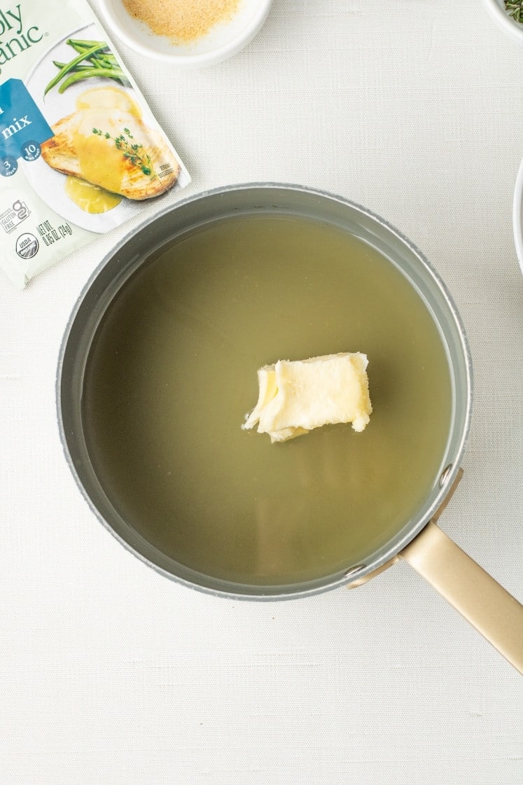 A saucepan of chicken broth with a pat of butter floating in the center, next to a seasoning packet.