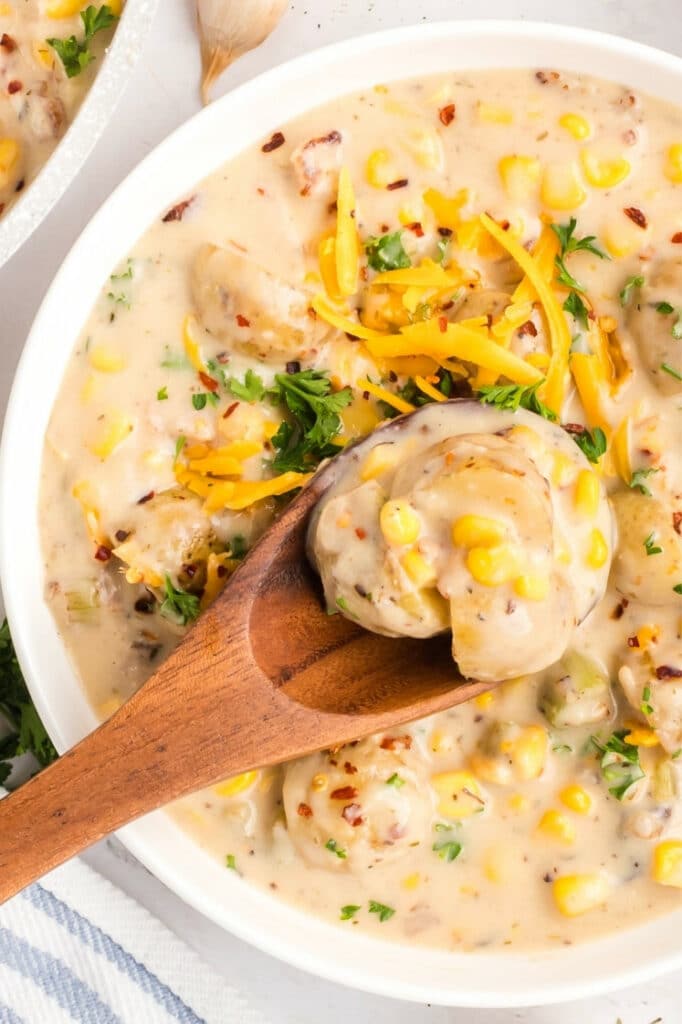 Close-up of a wooden spoon lifting baby potatoes from a bowl of creamy corn chowder topped with cheddar and parsley.