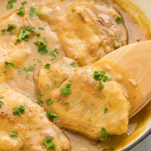 Close-up of a pork chop in honey mustard sauce on a wooden spoon, with more pork chops visible in the dutch oven below.