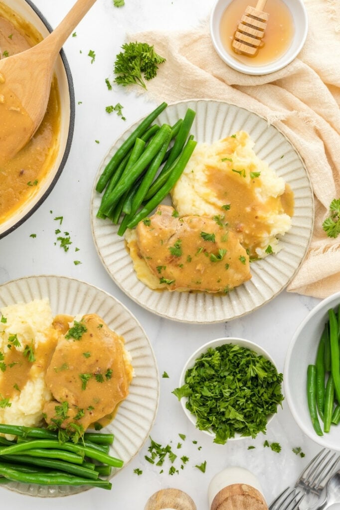 Overhead view of two plates of honey mustard pork chops with mashed potatoes and green beans, styled with a bowl of honey and fresh parsley.