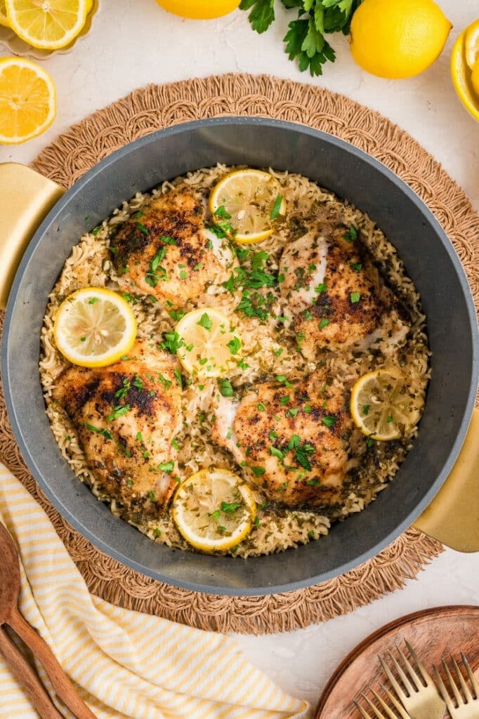 Overhead view of a dutch oven on a woven placemat filled with seared chicken thighs, lemon rice, and lemon slices, garnished with parsley.